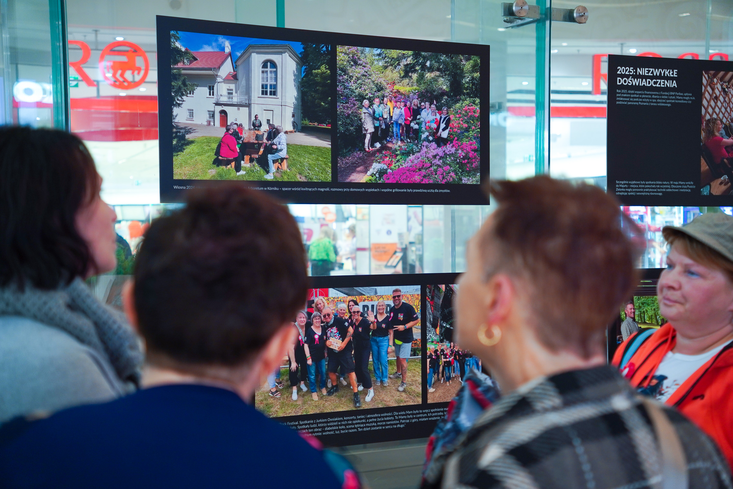 Kilka kobiet ogląda fotografie na planszach wystawy w galerii Posnania. Na zdjęciach widoczne są momenty z wcześniejszych spotkań projektu.
